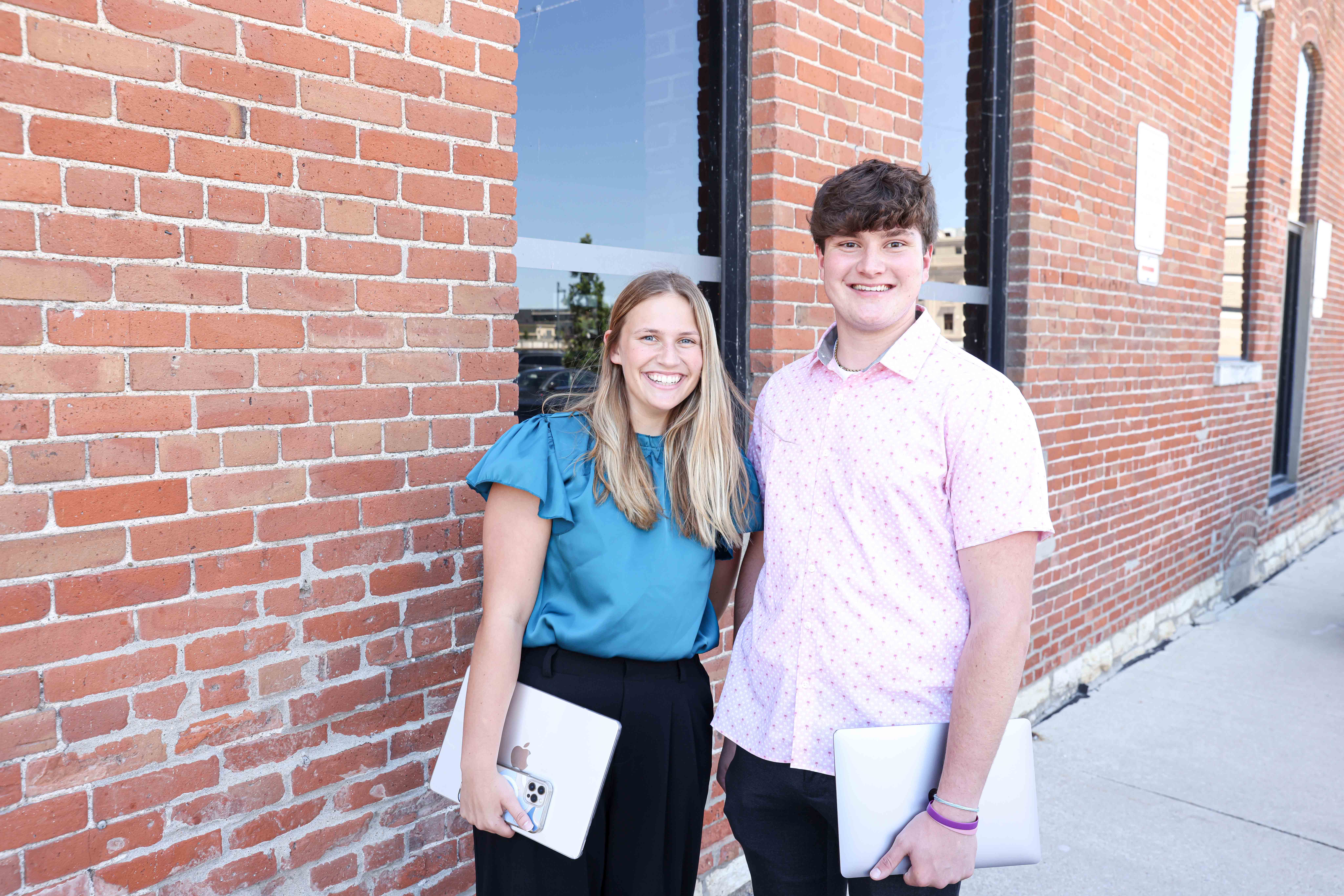 Two students smiling at the camera in front of a brick building where they report for their internship.
