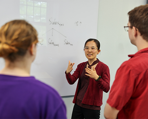 Professor Cai stands at a white board teaching two students.
