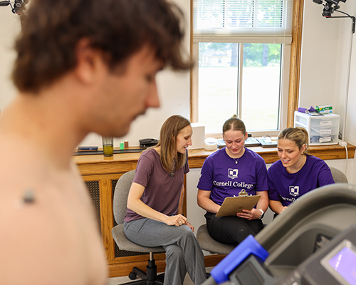 Two students and their professor discuss research, while a student runs on the treadmill in the foreground.