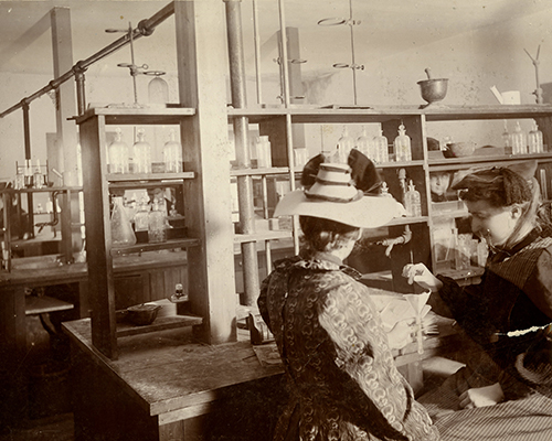 Two women conduct an experiment in a coed Cornell chemistry laboratory in 1898 (note the men visible behind them).
