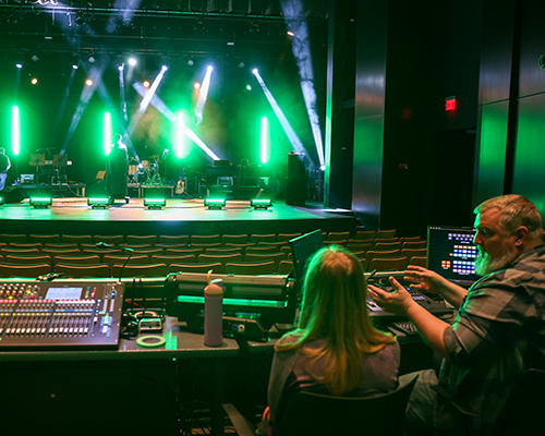 Scott Olinger teaches student lighting at a computer with the stage in front of them.