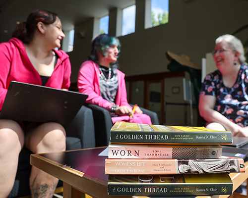 A stack of books that were read during CSRI in the foreground, with Professor of History Michelle Herder, Aslan Baker ’28, and Kat Maurelli ’27 in the background.