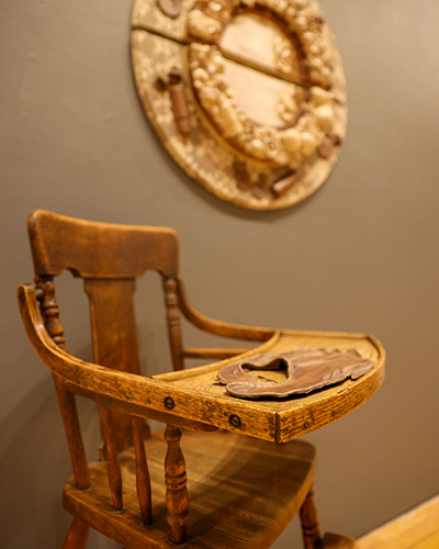 Art piece by Susannah that shows features a high chair, bib and another piece in the background.