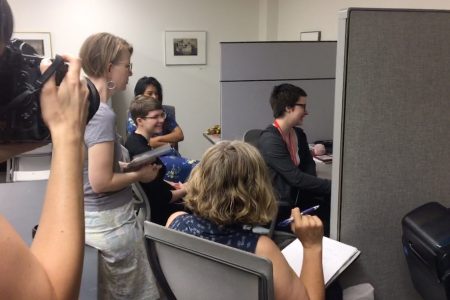 A photographer takes a picture of three students and two professors discussing a video playing on the computer monitor