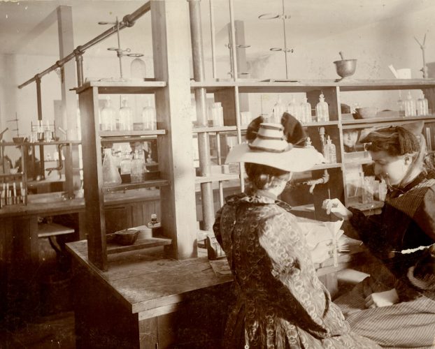 Two women conduct an experiment in a coed Cornell College chemistry laboratory in 1898 (note the men visible behind them). The woman at right is wearing an apron and oversleeves. 