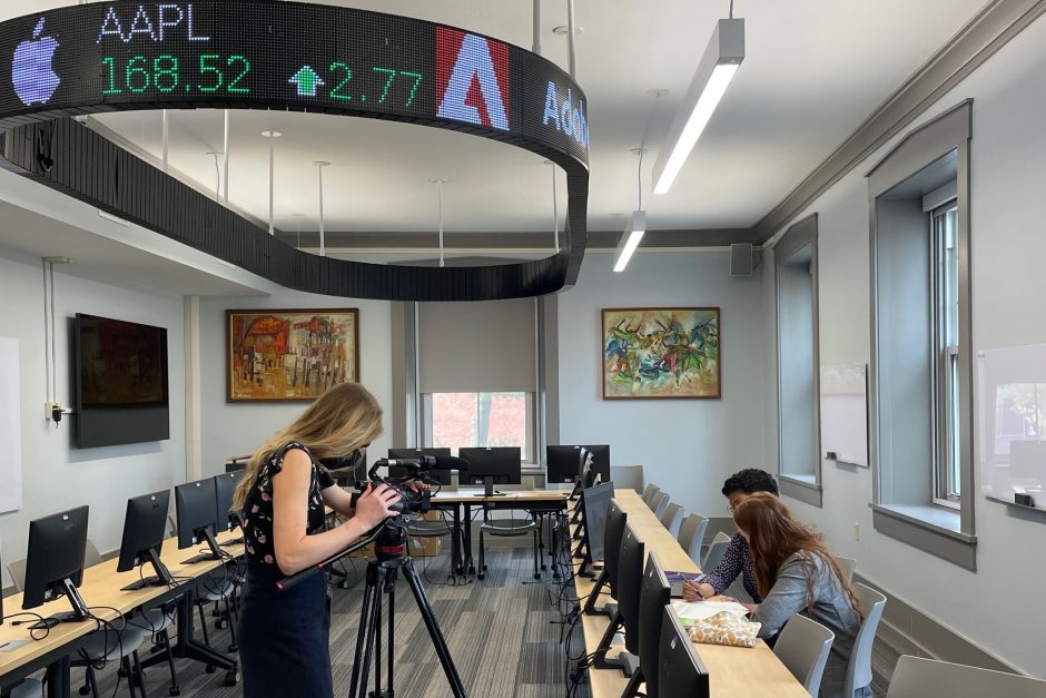 KCRG reporter takes video of Professor and student working at a computer in the finance lab with the financial ticker overhead