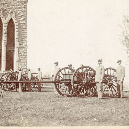 Uniformed men stand beside two breech-loading steel cannons in the late 18th century, with a portion of King Chapel visible behind them.