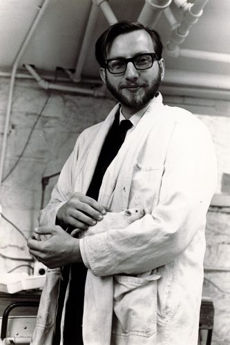 Black-and-white image of Professor Neil Wylie in a shirt, tie, and white lab coat, holding a white rat.