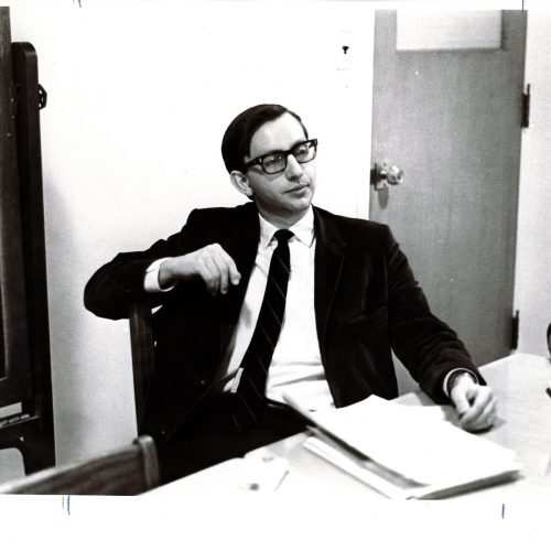 Black-and-white photo of Neil Wylie in suit and tie sitting at a table in a meeting.