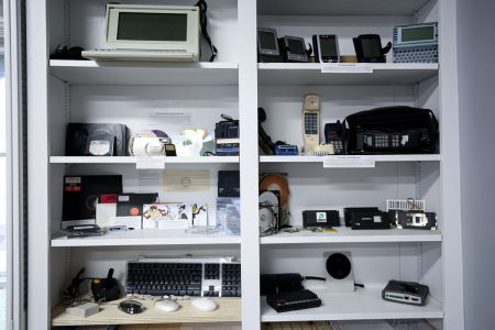 outdated technology including phones, computers, mice, keyboards, and storage is displayed on shelves in the Information Technology office