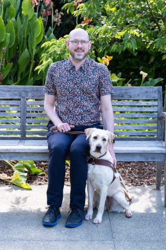 Shawn Henning '96 is seated on a bench outdoors with his golden Labrador guide dog seated on the ground next to him.