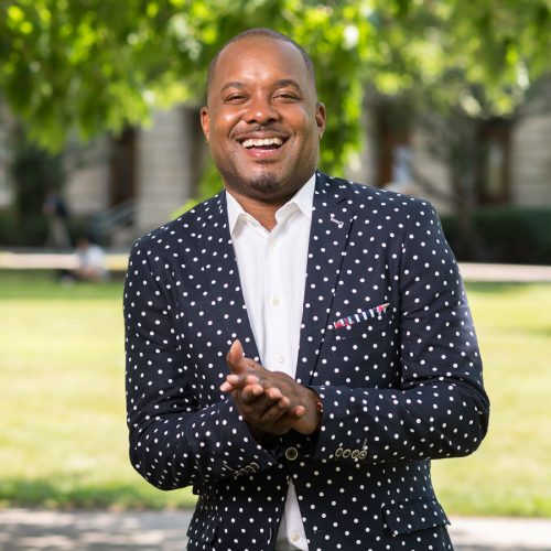 Jeffrey McCune standing on a campus in white shirt and black jacket with white polka-dots 