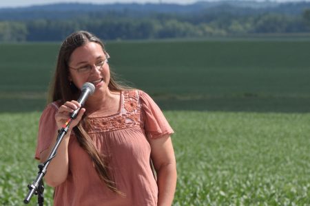 Jessica Wiskus with speaking into microphone with farm fields behind her