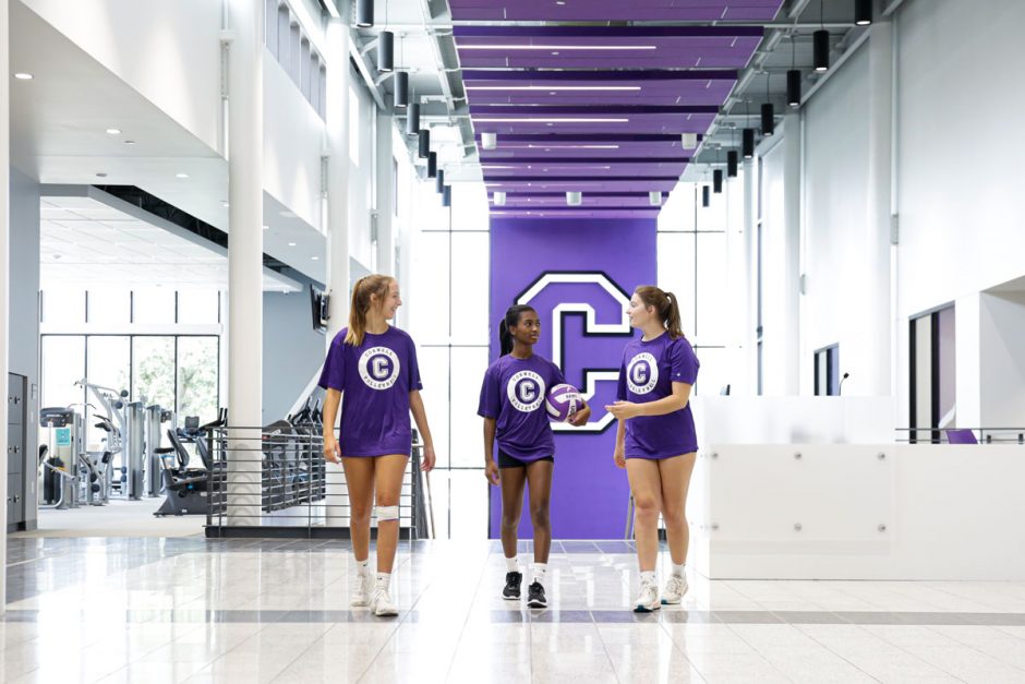 Volleyball players walk through the lobby of the SAW with the large athletic "C" logo on the wall behind them