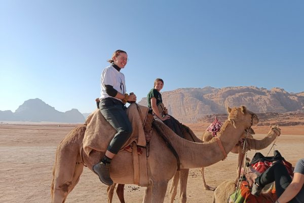Two students astride camels in a desert setting
