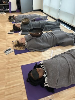 Four students laying on mats on the ground practicing yogic sleep with blankets and singing bowls