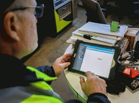 man wearing a fluorescent vest holding a tablet in an office space. There are file folders on the table as well as miscellaneous writing utensils