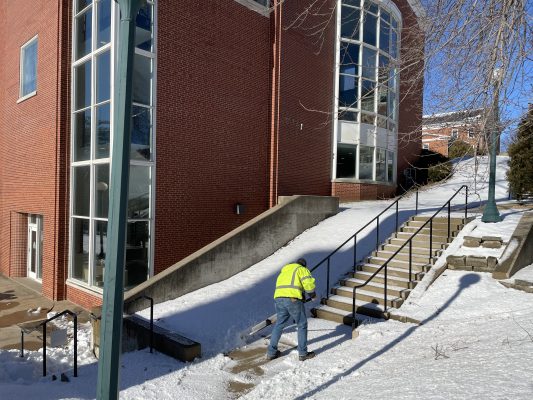 centered is a man wearing a fluorescent vest shoveling snow near a staircase. In the background there is Cole Library surrounded by snow 