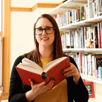 katie sagal holding a book open while smiling and bookshelves in the back