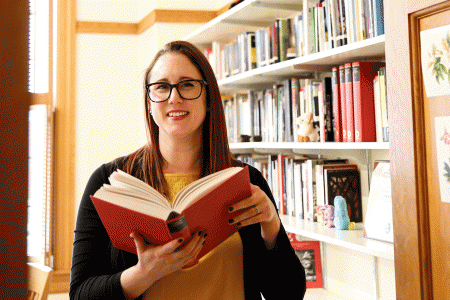 katie sagal is shown holding open a large hardcover book in front of a bookshelf full of books