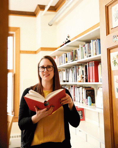 Katie sagal is shown holding an open hardcover book, in the backdrop is a bookshelf full of books