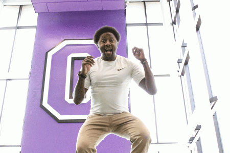 Lewis Kanyiba is jumping, he is completely in the air. The background is a purple sign and the cornell C