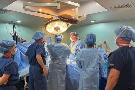 Several surgeons overlook a patient covered in a blue tarp with a bright light overhead.