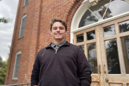 Senior Luke Baldwin stands in front of a red brick building on the Cornell College campus, smiling.