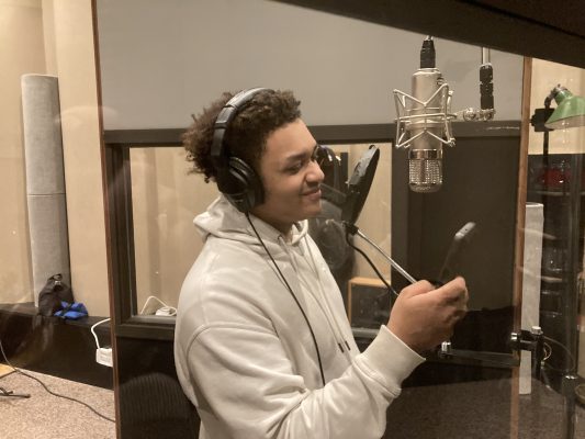 Student stands in in a professional recording studio space by a microphone and holding his phone.