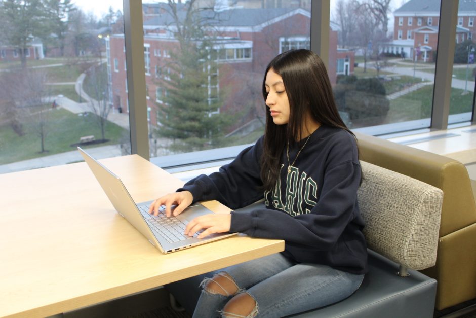 Natalia Alvarado Martinez working on a laptop in a lounge with windows and campus buildings in the background.