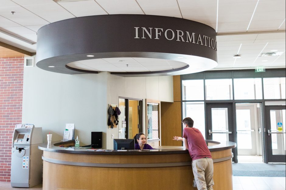 A student worker at the Information Desk talks to a student leaning on the round desk.