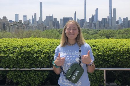 Indy Smith stands holding two thumbs up with a city skyline in the background.