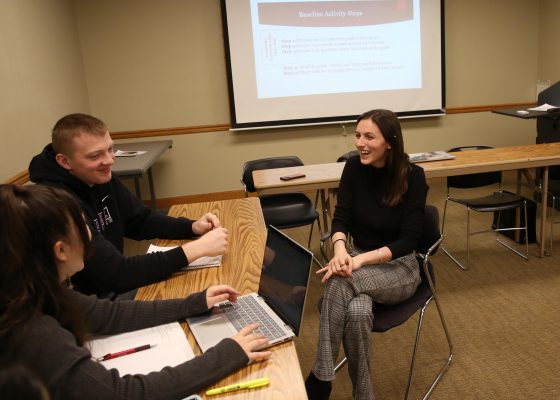 Instructor sitting across from two students, smiling as they discuss their work.