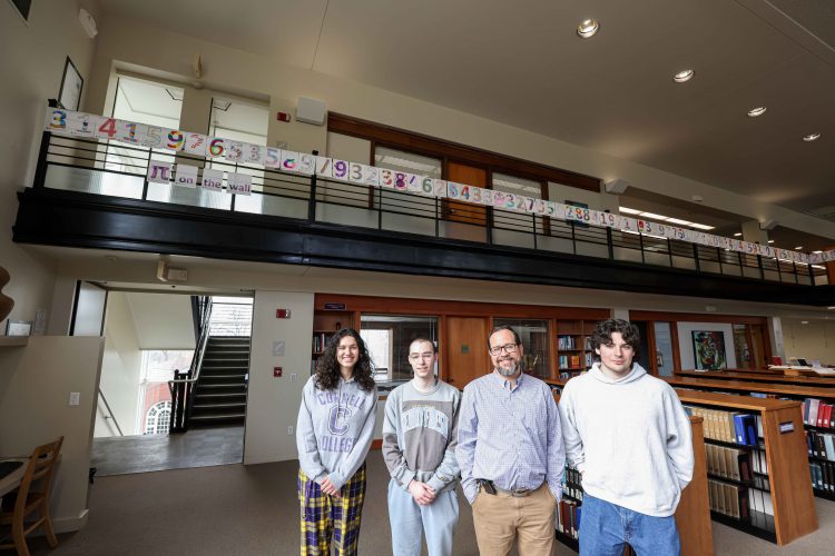 Three students and a professor stand in Cole Library with digits of Pi above them on the balcony.