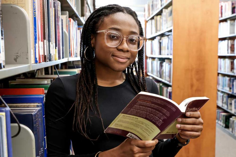 Kimberly Maitland '24 peers over an open book she's holding among the stacks in Cole Library.