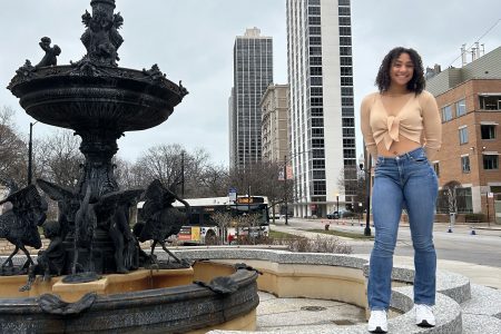 Cotton stands by a fountain with a city skyscape in the background.