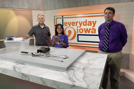 Sarah Renaud and two professors, Joshua Otten and Tyler George, stand behind a set on Everyday Iowa at KCRG in downtown Cedar Rapids. Sarah is holding a turtle.