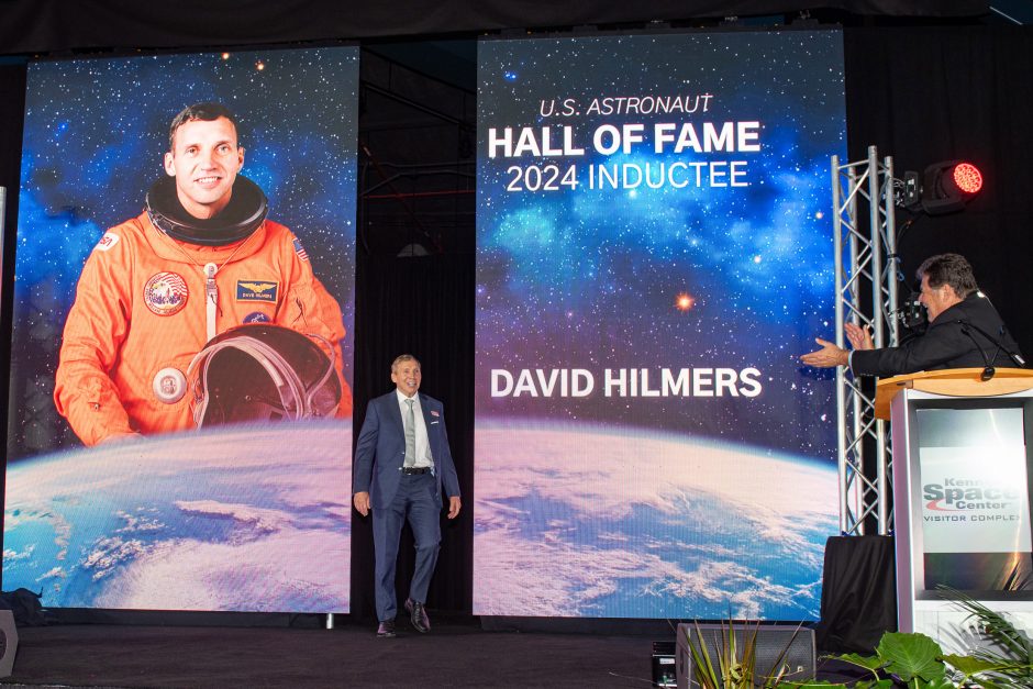 David Hilmers walking toward a podium on a stage with large banners of his photo and his name.