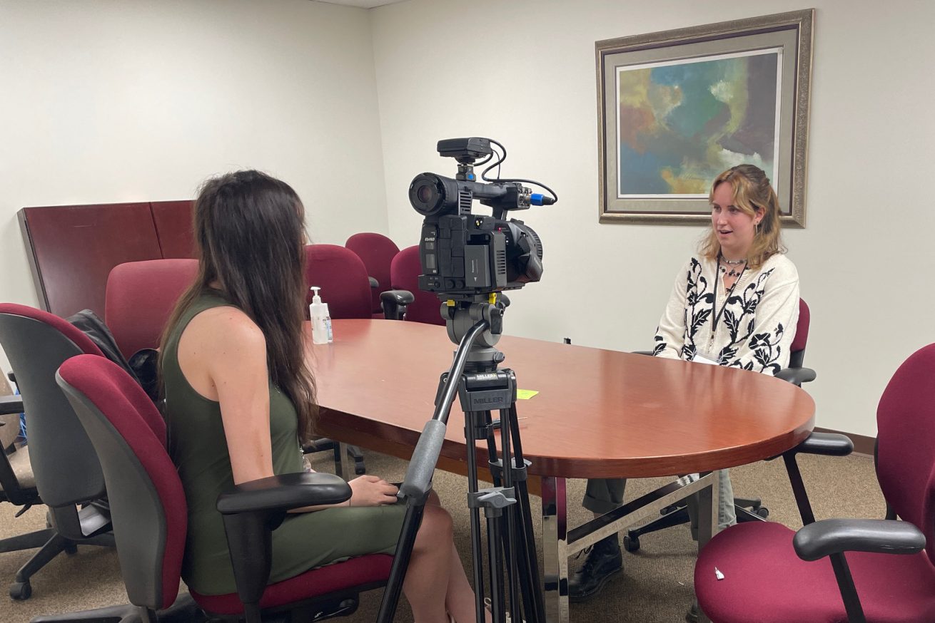 Reporter sits behind camera and asks Oriana questions on camera in a conference room.