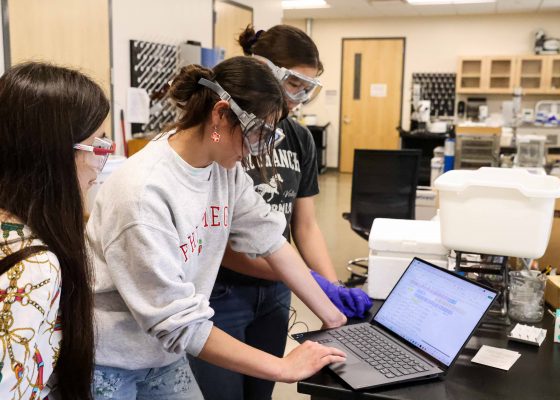 two students and their professor looking at graphs and data on a laptop in a lab setting.