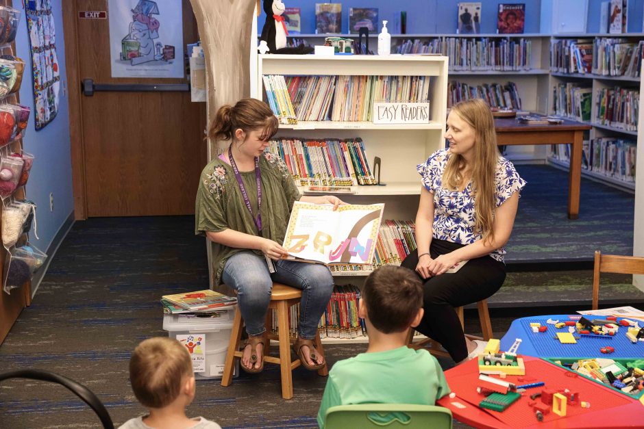 Nellie sits next to librarian and reads a book to local children.