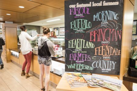 Two students get served food and in the foreground there's a sign with the names of several local farms.