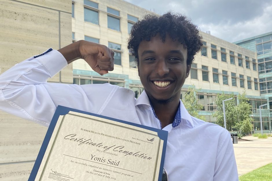 Yonis Said holding up an arm in a muscle pose as he holds his certificate of completion in the other hand.