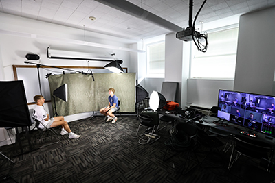 Two students sit in front of cameras with photographic lighting.