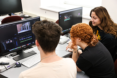 Three students sit around computer monitors with editing software on the monitor.