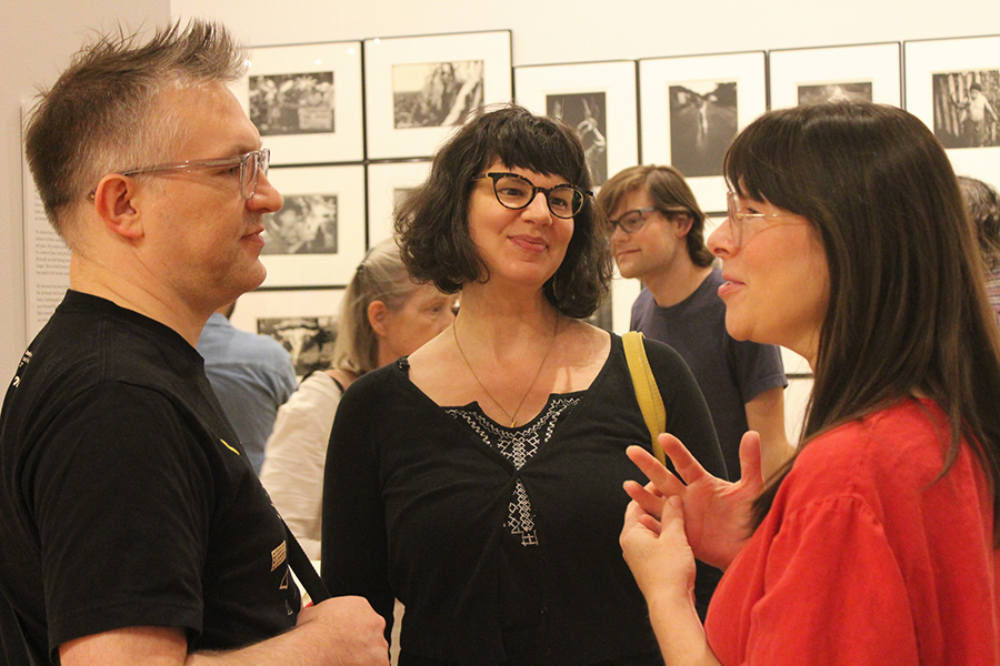 Three people talking with photos from an exhibit on the wall behind them.