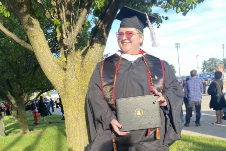 Finley Cowlishaw ’19 in academic regalia holds their master's degree diploma